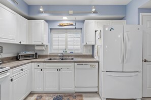 A kitchen with white cabinets, a built-in dishwasher, a refrigerator, and a window with blinds. - 🏖️Spacious 3BR Retreat: Pool, Tennis & Estuary Views | Oceanwalk 7-207🦈 (New Smyrna Beach)