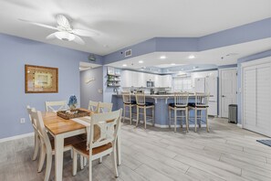 A modern kitchen with white cabinets, a central island with bar stools, and a dining area with a wooden table and chairs. - 🏖️Spacious 3BR Retreat: Pool, Tennis & Estuary Views | Oceanwalk 7-207🦈 (New Smyrna Beach)