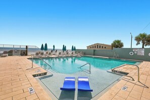 A swimming pool with lounge chairs and umbrellas, a fenced area, and a building in the background. - 🏁Daytona Beach Club 614 | Oceanfront Complex + Pool 🌴 (Daytona Beach)