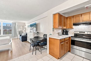 A modern kitchen with wooden cabinets, a stainless steel oven, and a dining area with a table and chairs. - 🏁Daytona Beach Club 614 | Oceanfront Complex + Pool 🌴 (Daytona Beach)