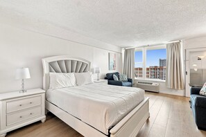 A hotel room with a large bed, a nightstand, a chair, a window with a view of buildings, and a door. - 🏁Daytona Beach Club 614 | Oceanfront Complex + Pool 🌴 (Daytona Beach)