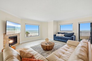 A spacious living room with a fireplace, a large sofa, a coffee table, and a view of the beach through large windows. - Seascape Towers 222: Beachfront Getaway with Balcony & Heated Pool 🏖️ (New Smyrna Beach)