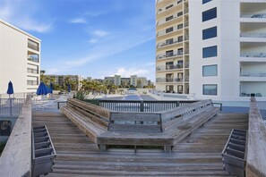 A wooden boardwalk leading to a pool area with lounge chairs and umbrellas, surrounded by high-rise buildings. - Seascape Towers 222: Beachfront Getaway with Balcony & Heated Pool 🏖️ (New Smyrna Beach)