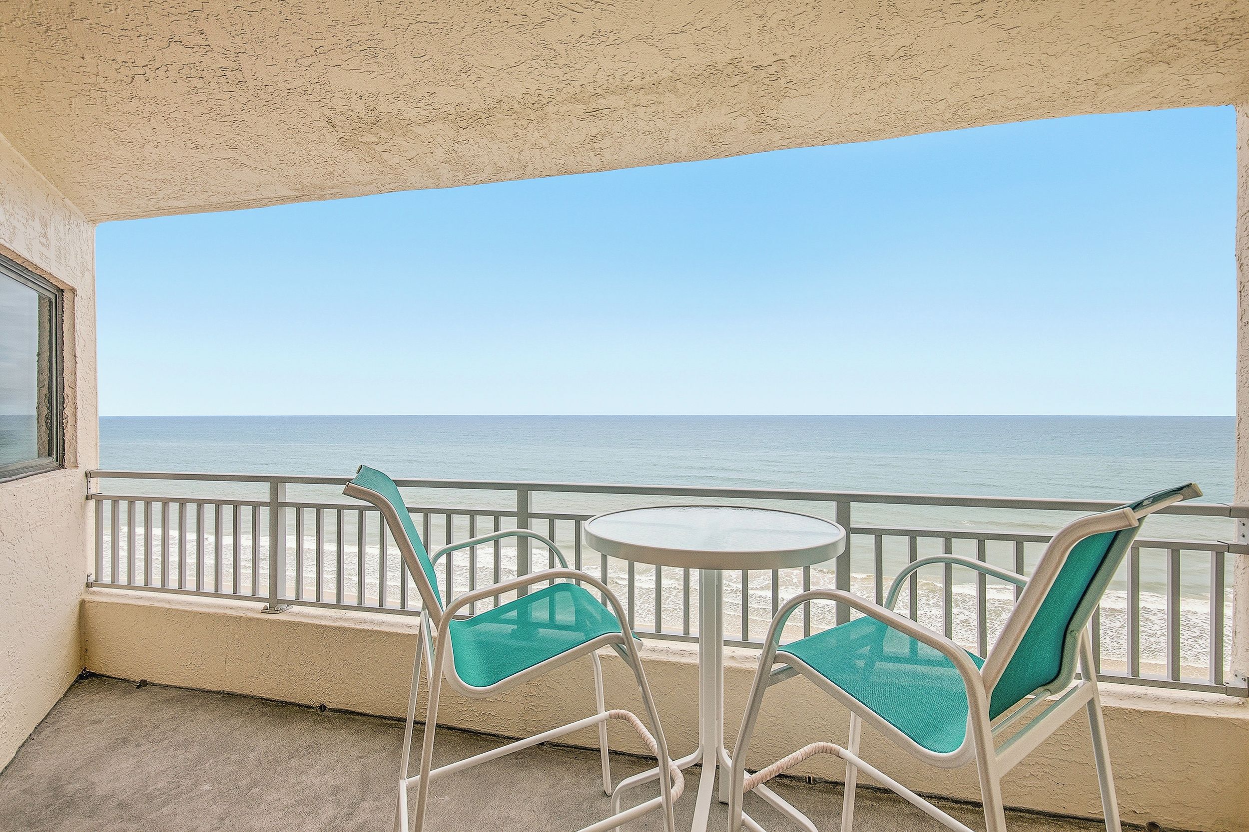 A balcony with a view of the beach and ocean. There are two chairs and a small round table.