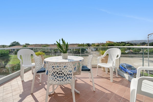 A terrace with a round table and white chairs, a potted plant, and a view of a blue car and distant buildings.