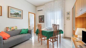 A living room with a sofa, a dining table with chairs, a television, and a window with curtains.
