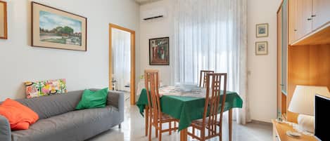 A living room with a sofa, a dining table with chairs, a television, and a window with curtains.