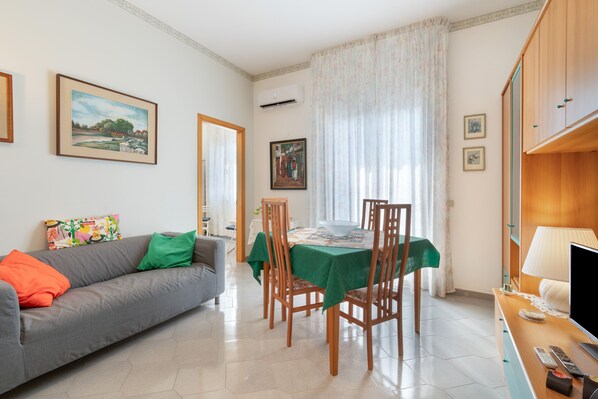 A living room with a sofa, a dining table with chairs, a television, and a window with curtains.