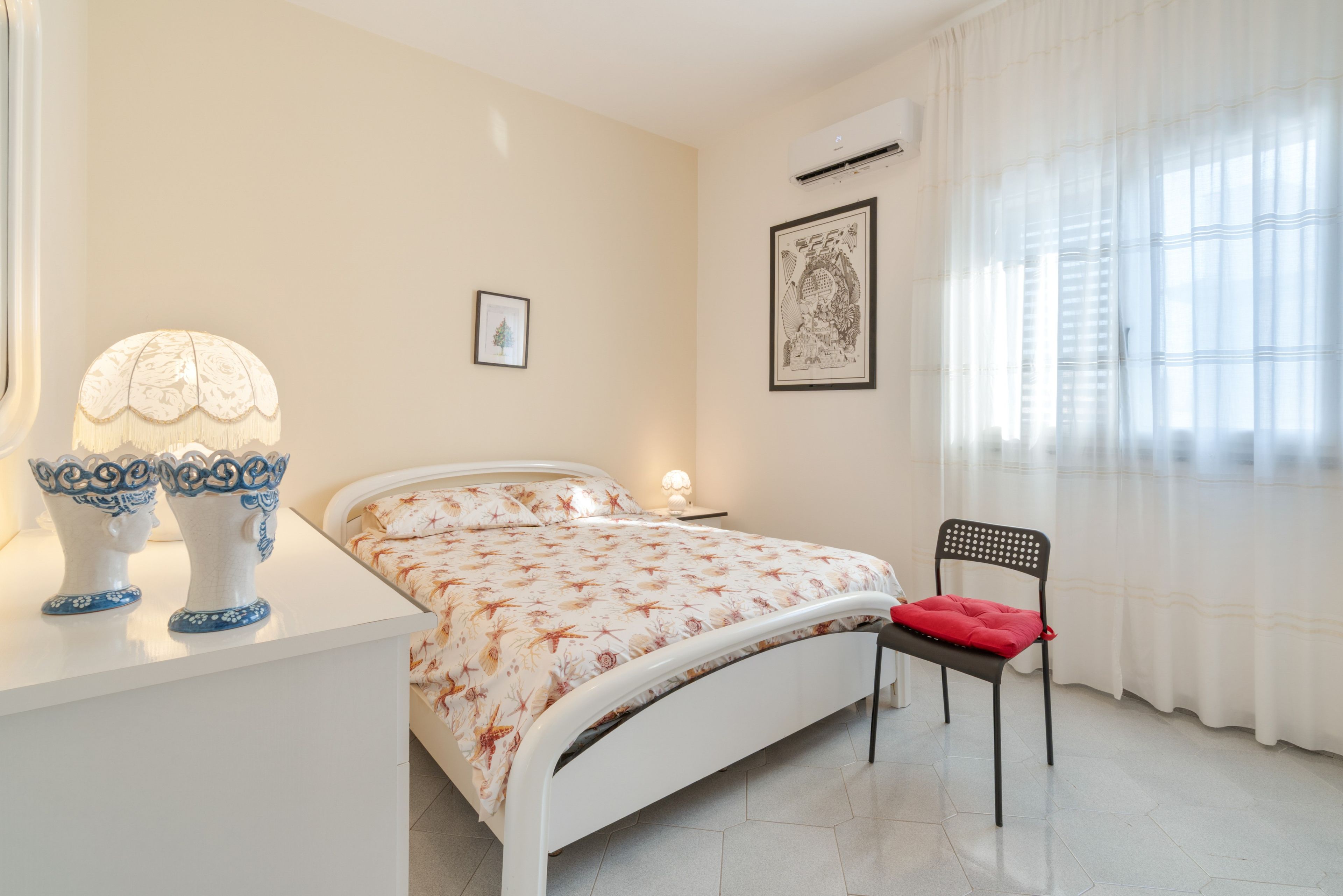 Un dormitorio con cama, mesita de noche, lámpara, silla y una ventana con cortinas.