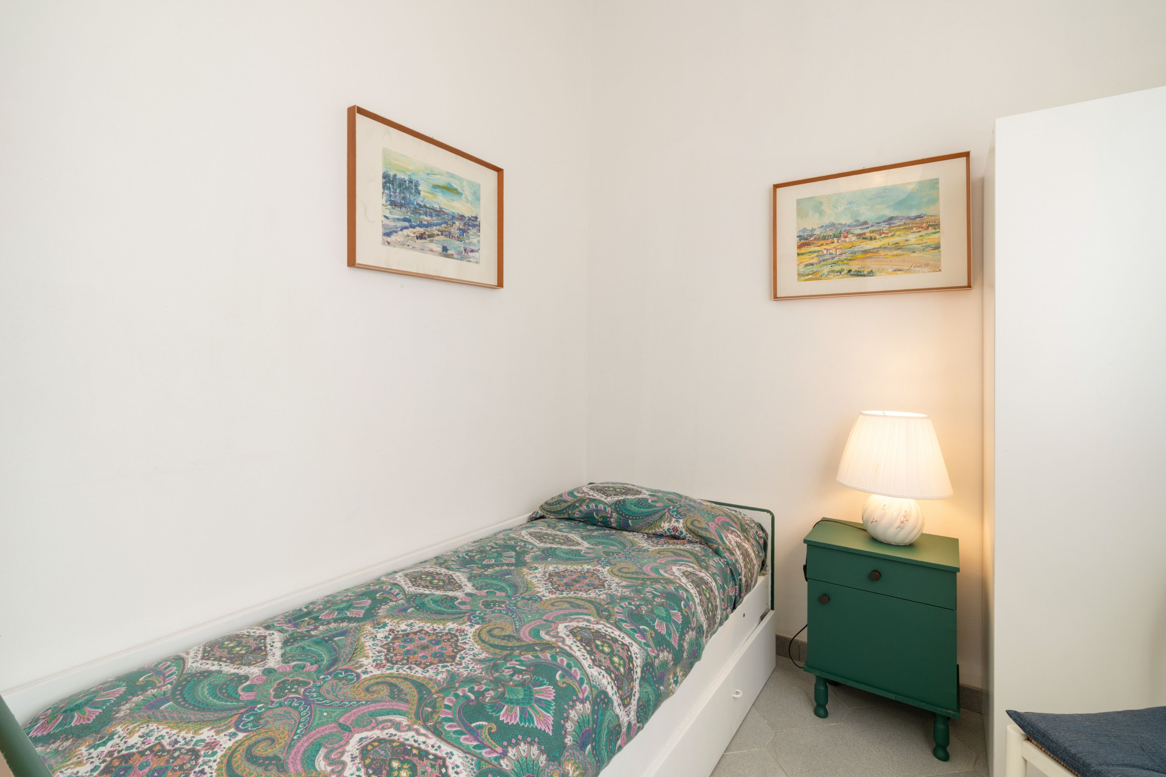 A small, single-bed room with a green nightstand and a lamp, two framed paintings on the wall, and a white door.