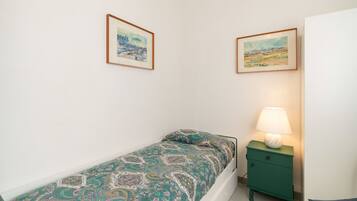 A small, single-bed room with a green nightstand and a lamp, two framed paintings on the wall, and a white door.