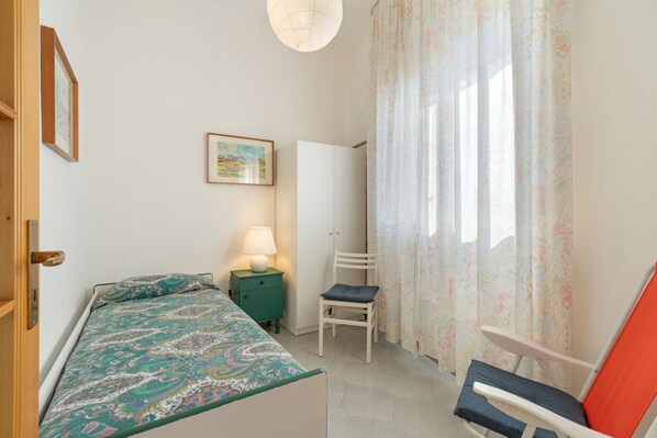 Un dormitorio con cama, una silla, una mesita de noche con lámpara, un cuadro en la pared y una ventana con cortinas.