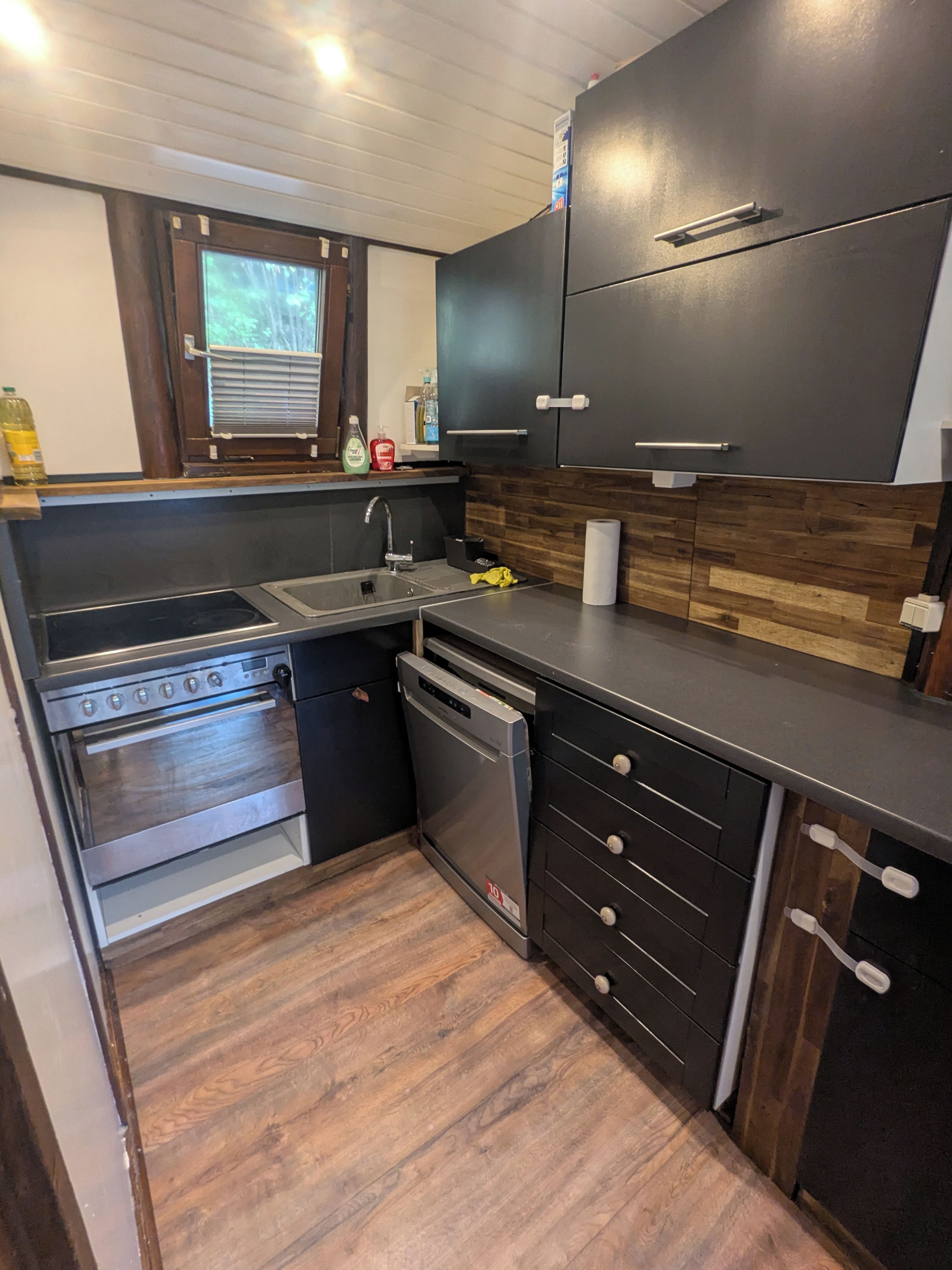Eine kleine Küche mit eingebautem Backofen, Geschirrspüler und einer Holzrückwand.