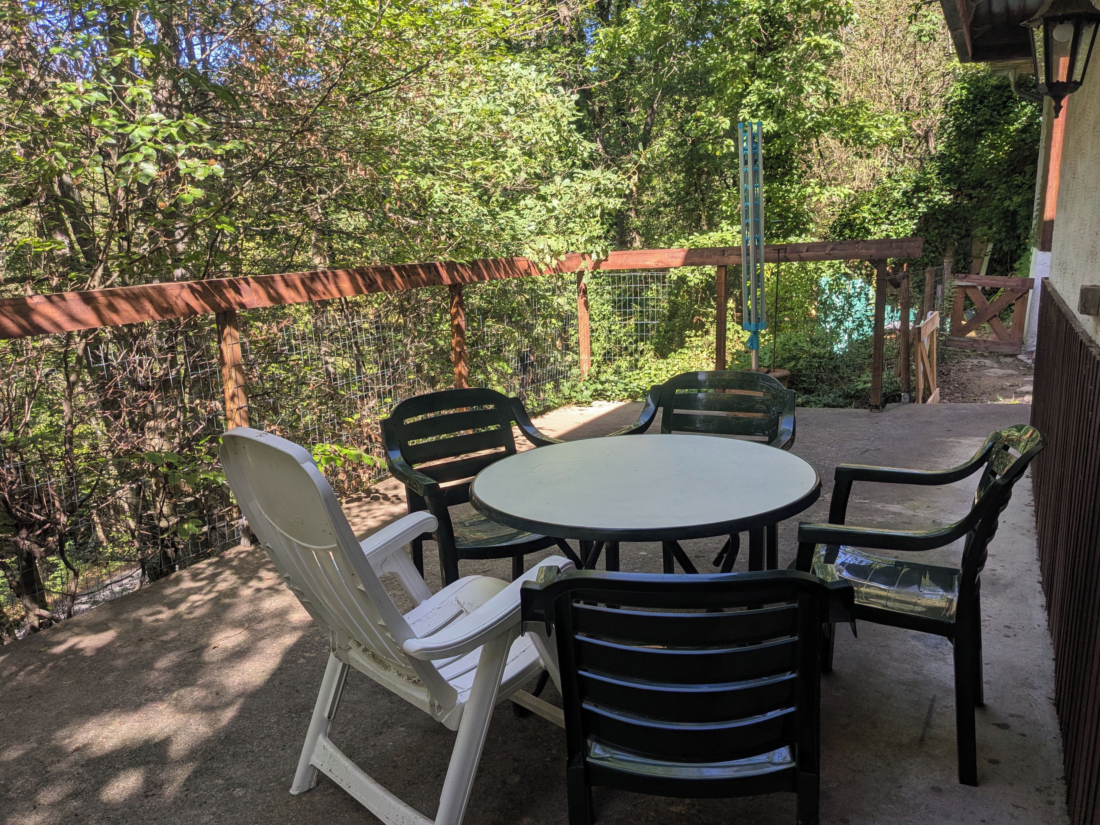 En udendørs terrasse med et rundt bord og stole, omgivet af et træhegn og grønt.