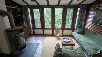 A cozy cabin interior with a wood-burning stove, a green sofa, a wicker chair, and large windows offering a view of the forest.