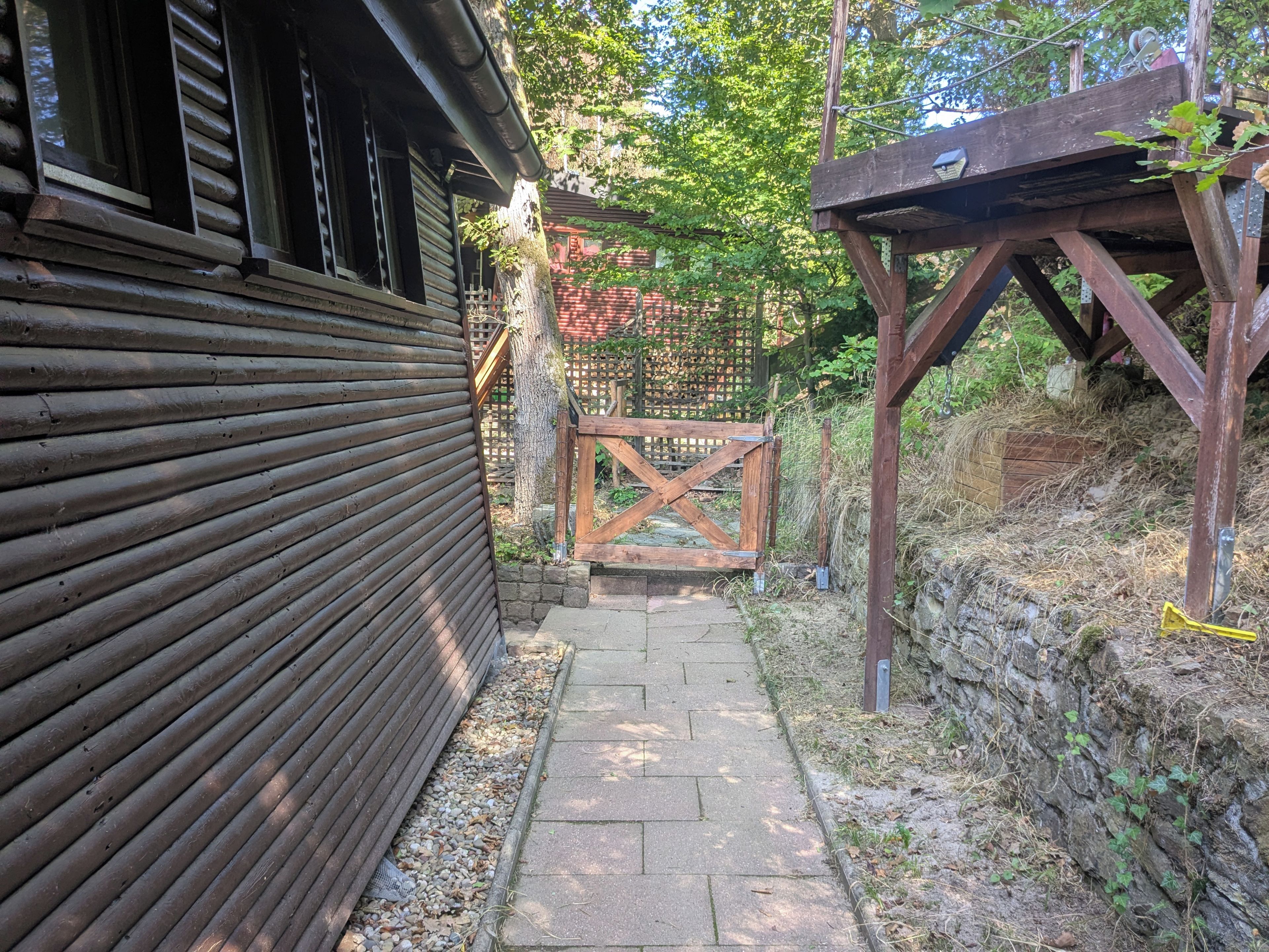 Ein schmaler Steinweg führt zu einem Holztor, begleitet von einem Gebäude mit Holzverkleidung und einer hölzernen Pergola.