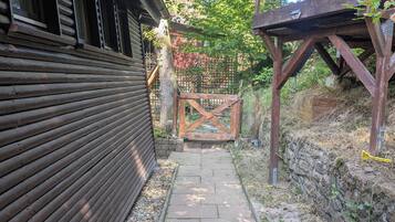 Uma estreita trilha de pedra leva a um portão de madeira, ladeada por um prédio de madeira com revestimento horizontal e uma estrutura de pérgola.