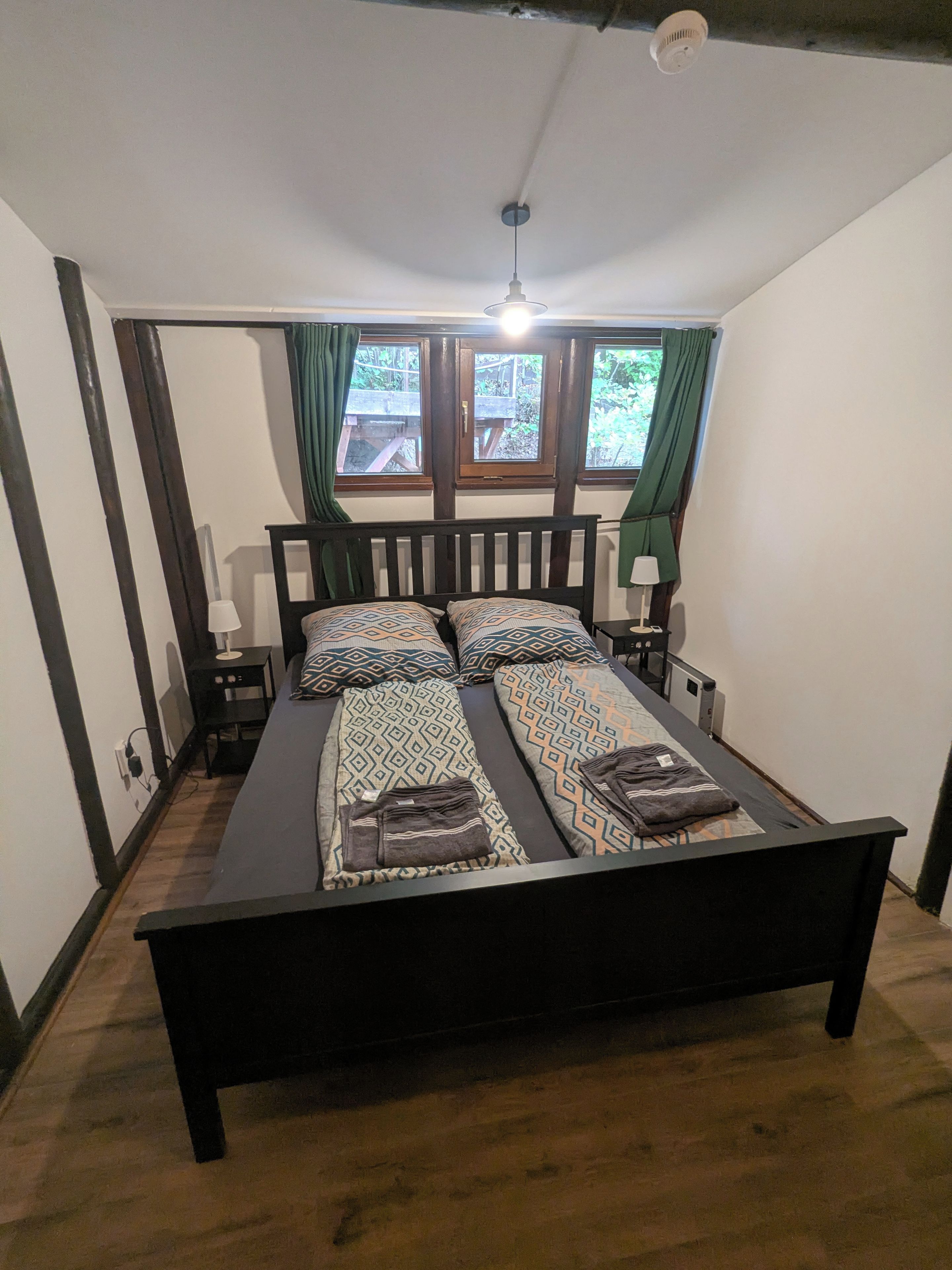 En dobbeltseng med mønstret sengetøj, et sengebord med lampe og et vindue med gardiner.