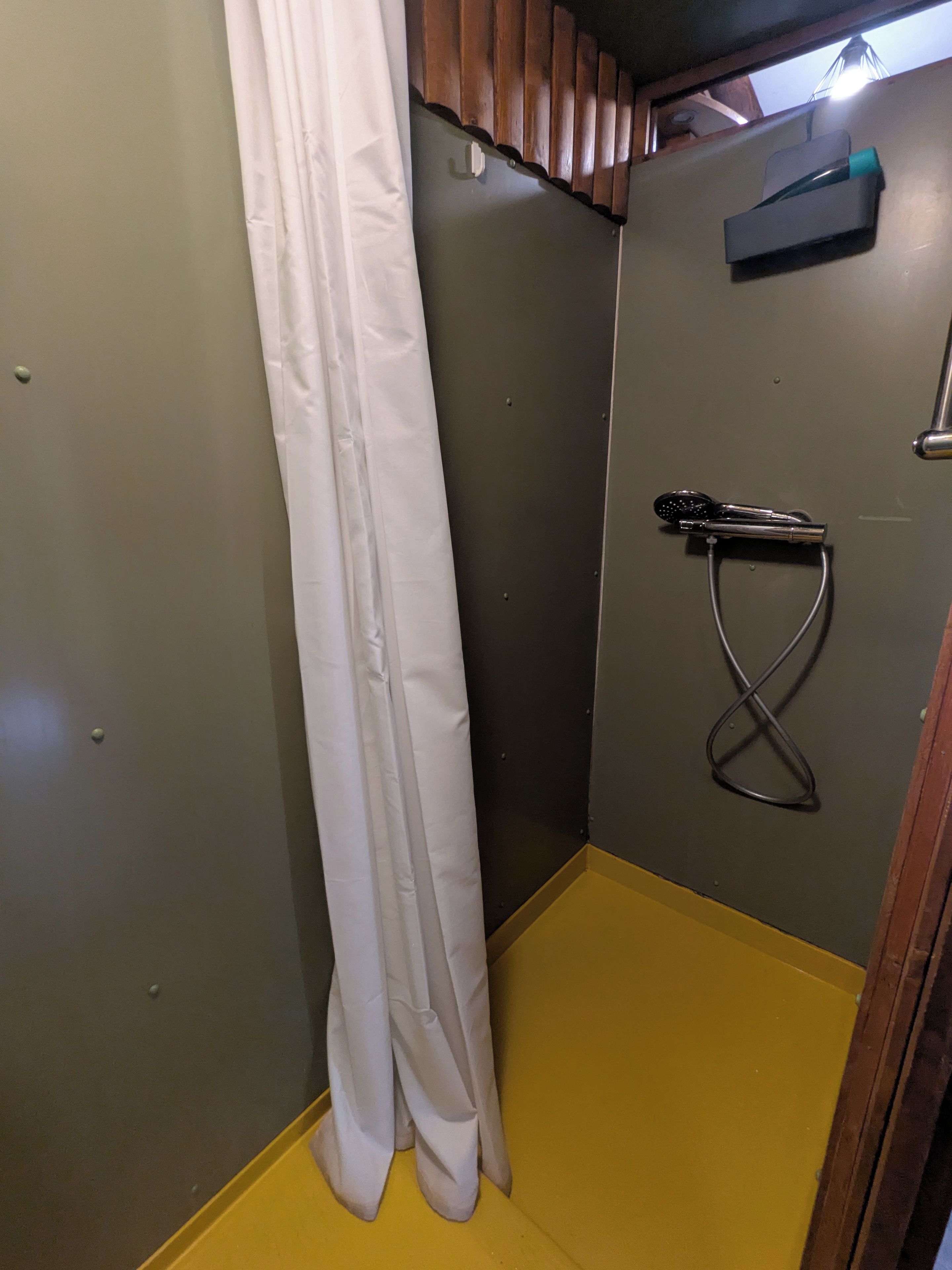 Un área de ducha pequeña y cerrada, con piso amarillo, cortina de ducha blanca y un cabezal de ducha de mano.