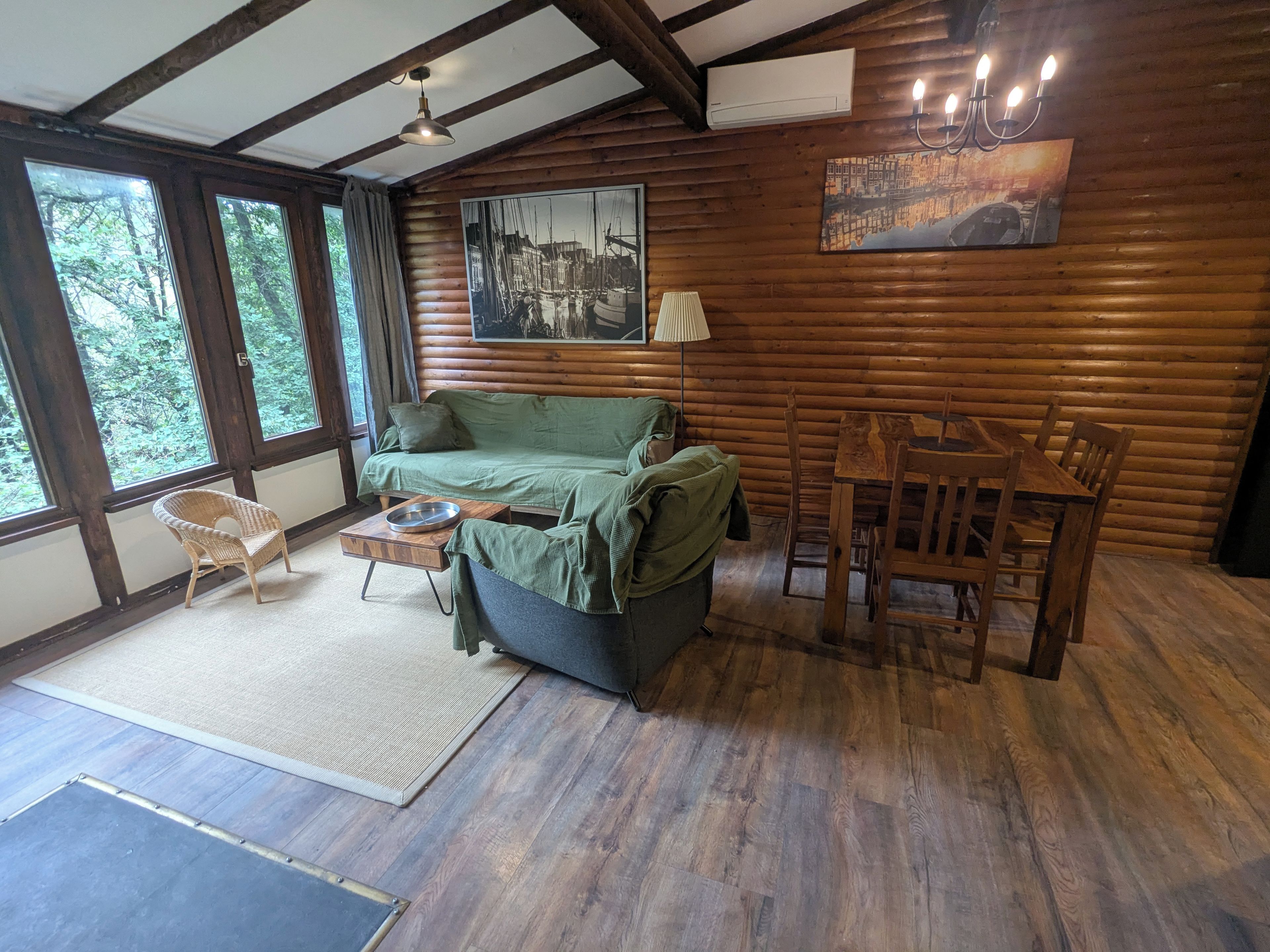 L’intérieur chaleureux d’un chalet avec un plancher en bois, un canapé vert, une table à manger avec des chaises et de grandes fenêtres donnant sur la forêt.
