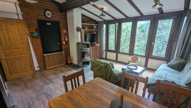 Een gezellige kamer met een houten tafel, stoelen, een bank, een open haard en grote ramen met uitzicht op de omgeving.