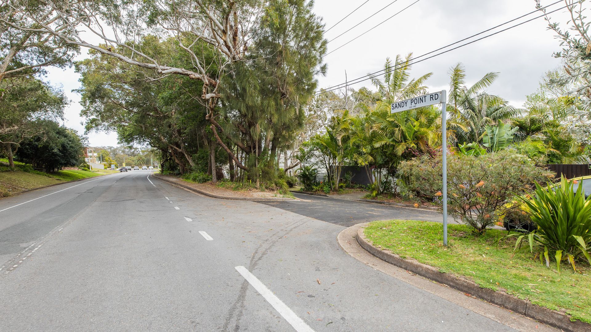 A street view with a road marked 'SANDY POINT RD.' There are trees and a sidewalk on the right, and a building with a fence in the background.