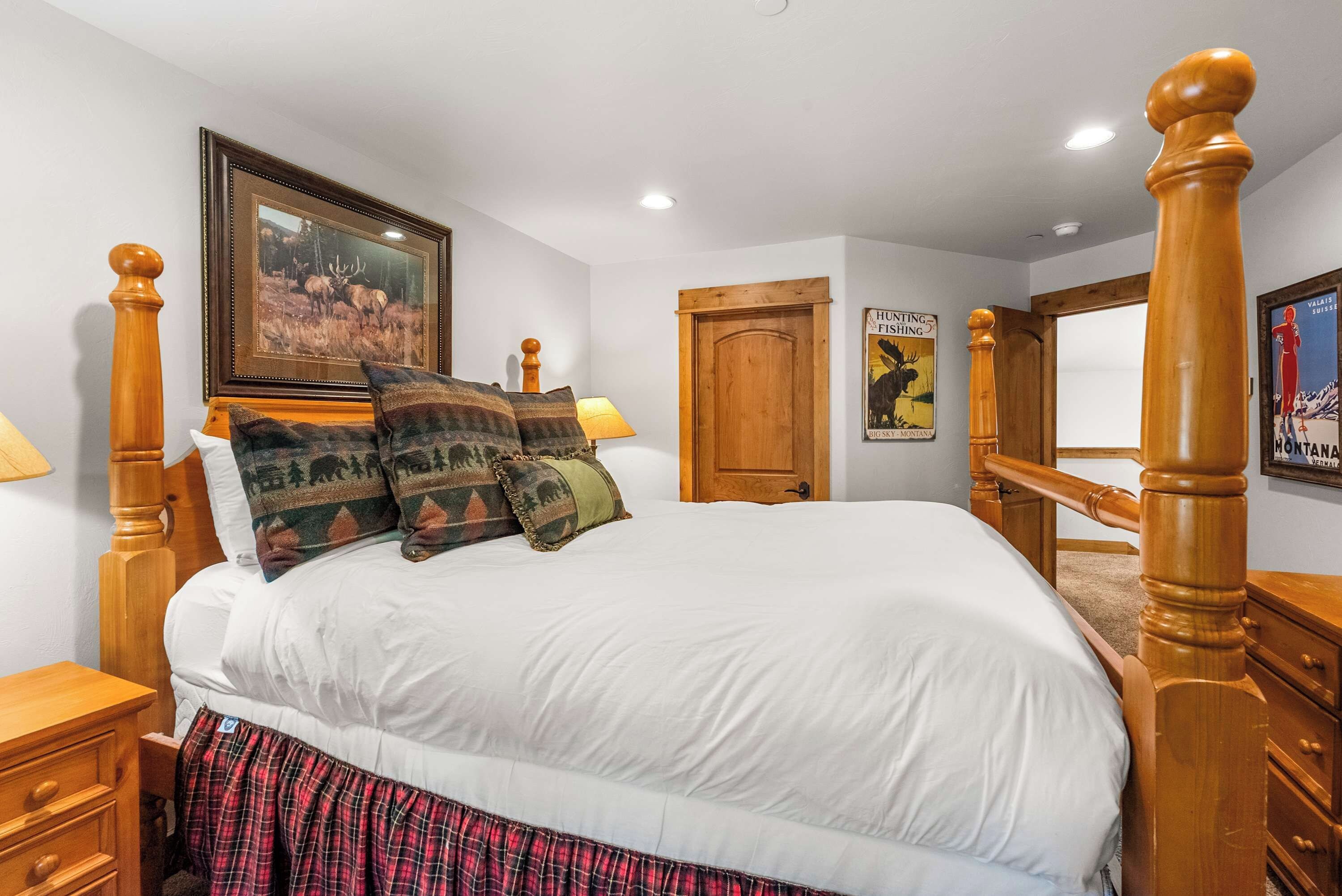 A bedroom with a large bed, wooden headboard, a framed picture of elk, and a door with a poster.