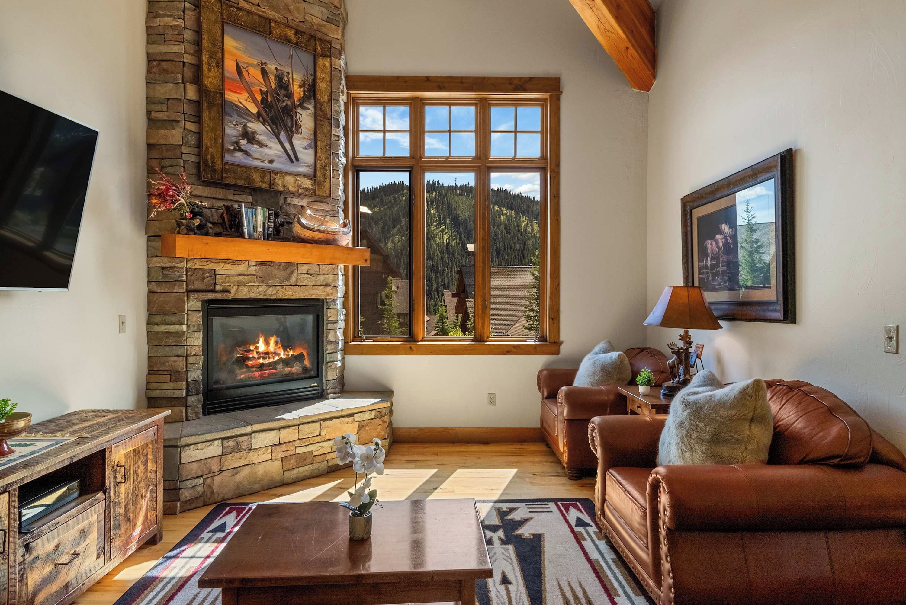 A cozy living room with a stone fireplace, leather sofas, a flat-screen TV, and a large window with a mountain view.