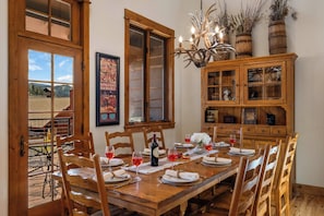 A dining room with a wooden table set for six, surrounded by chairs. There is a chandelier with antlers, a wooden cabinet with glass doors, and a framed poster on the wall. - Big Sky Gem - Mtn Views + Private Deck + Hot Tub (Gallatin Gateway)