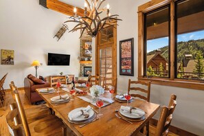 A dining room with a wooden table set for a meal, a fireplace, and a view of a cabin through a large window. - Big Sky Gem - Mtn Views + Private Deck + Hot Tub (Gallatin Gateway)