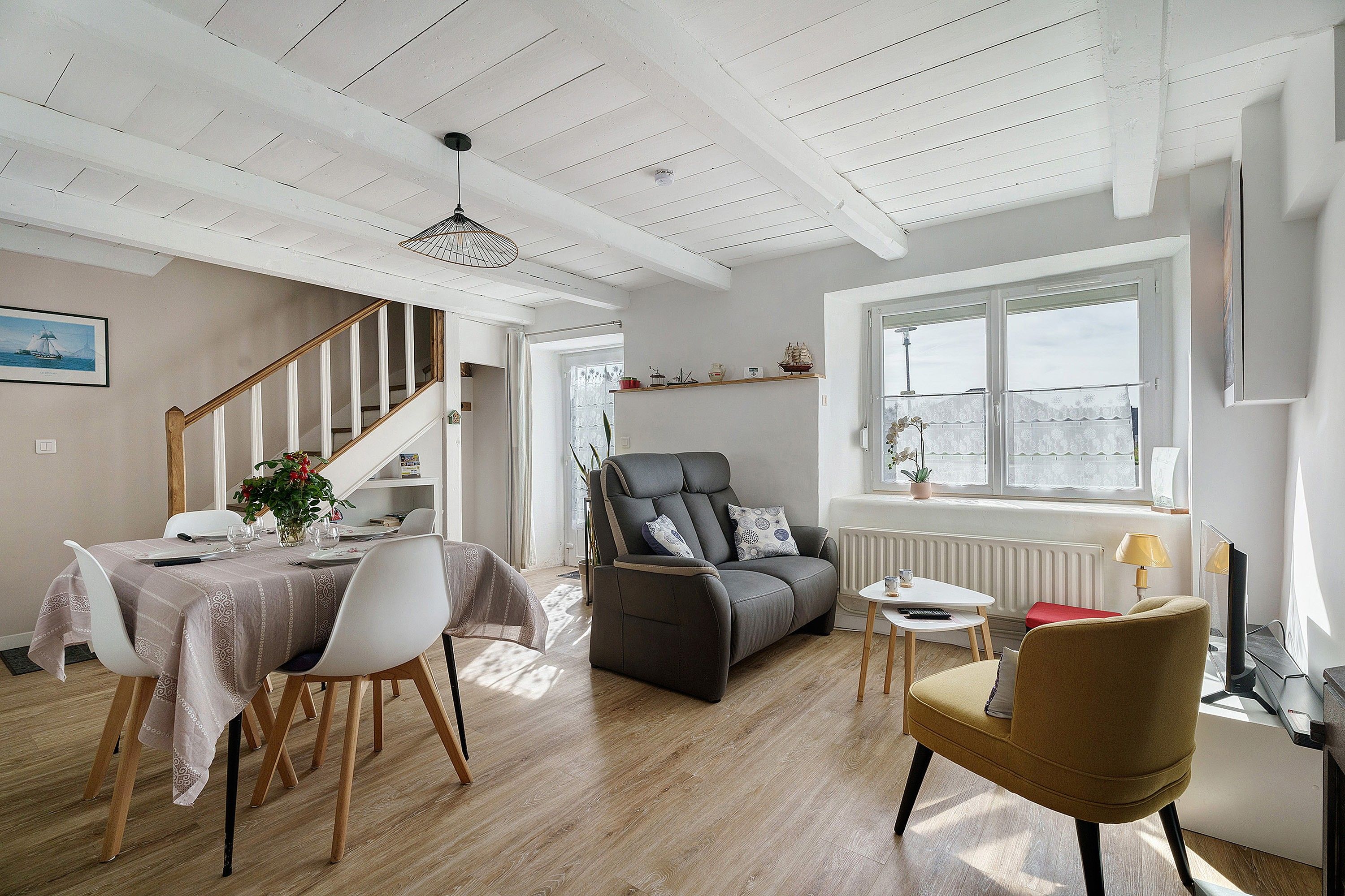 A spacious living area with a dining table set for four, a grey sofa, a yellow armchair, and a window with a view.