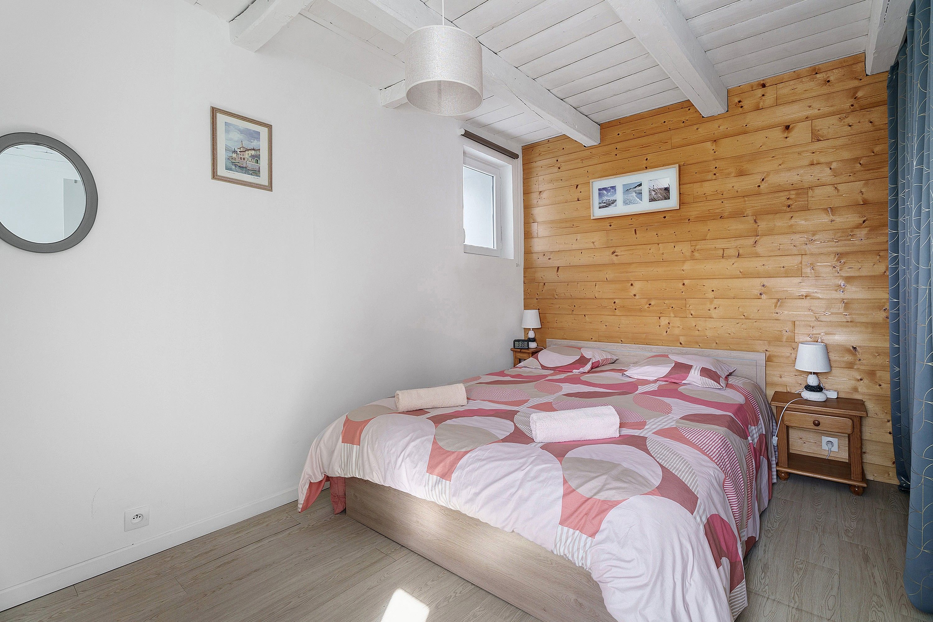 A bedroom with a wooden headboard, a bed with a patterned bedspread, two bedside tables with lamps, a mirror on the wall, and a window.