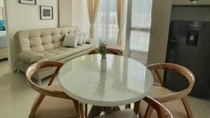 Un salon avec un canapé, une table à manger avec un plateau en verre et des chaises, un coin cuisine avec un réfrigérateur, et un balcon avec des rideaux.