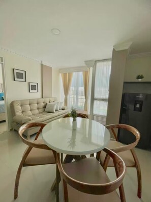 Sala de estar com sofá, mesa de jantar com tampo de vidro e cadeiras, cozinha com geladeira e varanda com cortinas.