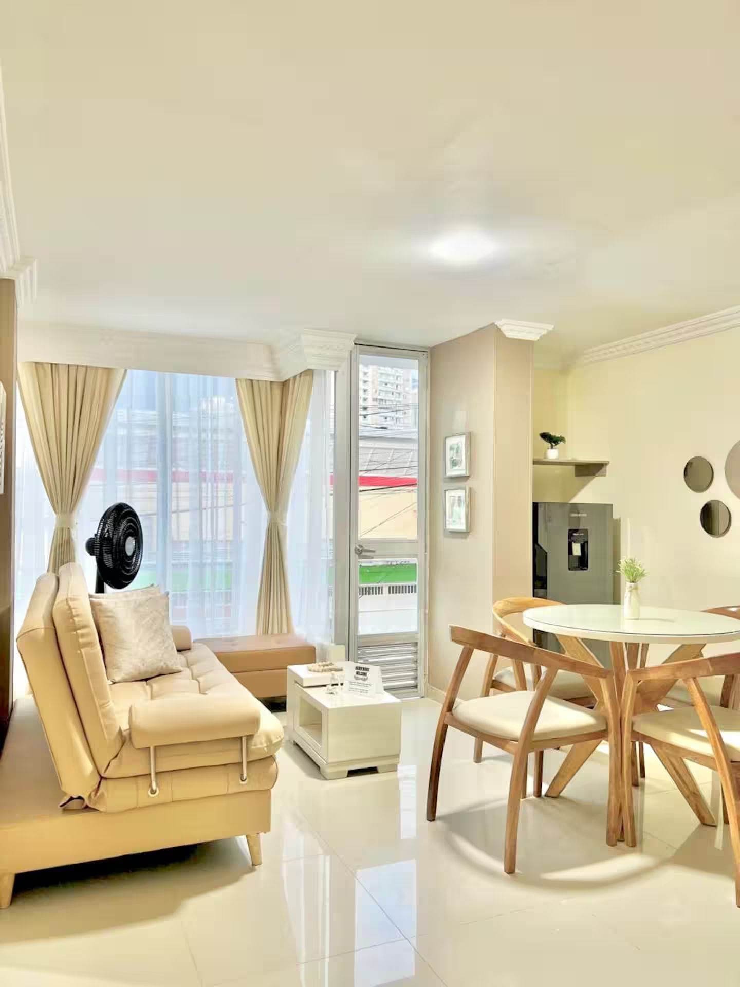 Una sala de estar moderna con un sofá, una mesa de comedor con sillas, un ventilador y vistas a la ciudad a través de amplios ventanales.