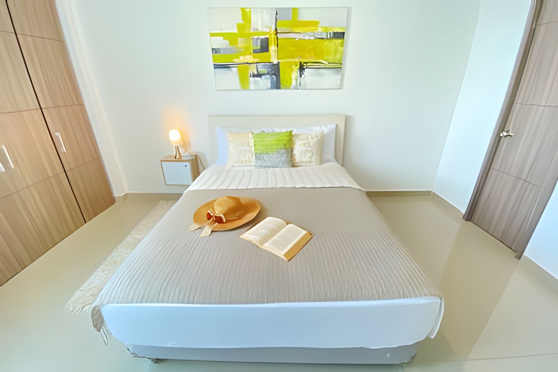 A neatly arranged bedroom with a bed, a bedside table with a lamp, and a wall-mounted abstract painting.