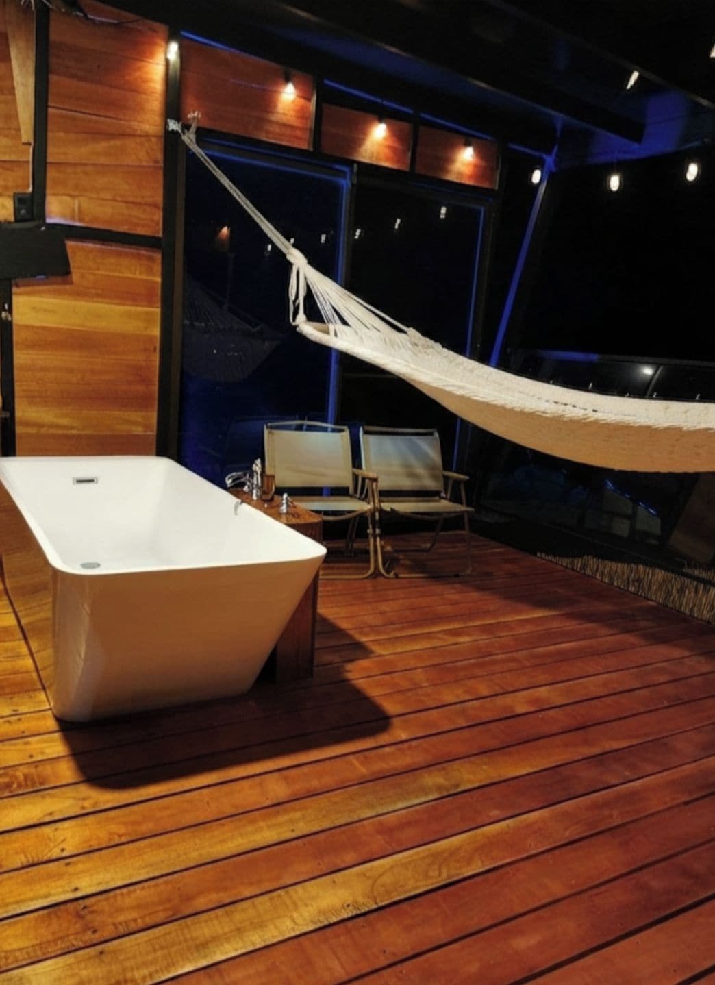 A hammock, a white bathtub, and two chairs on a wooden deck.