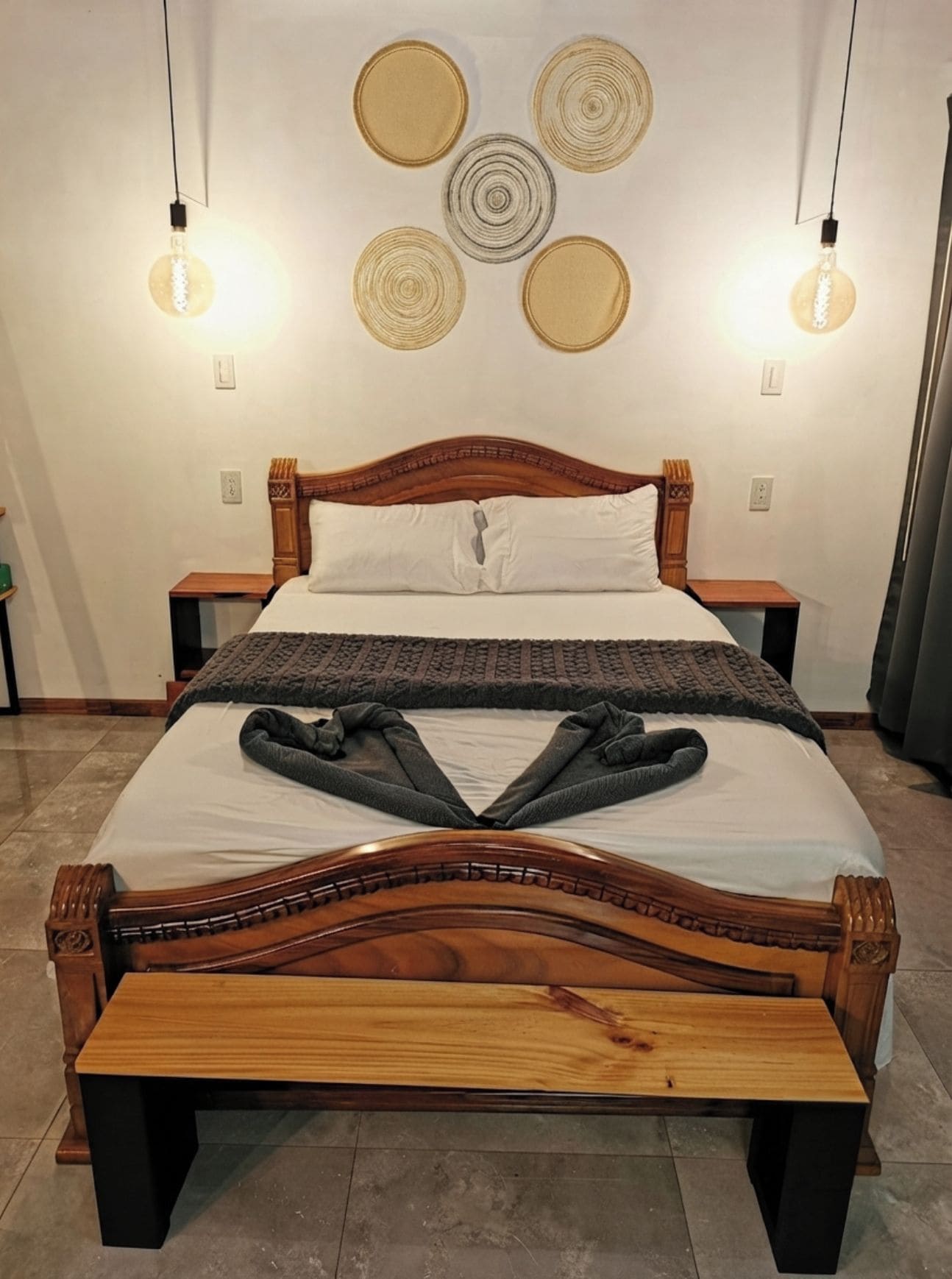 Un lit avec un cadre en bois, un matelas blanc et une couverture grise. Il y a deux tables de chevet avec des lampes.