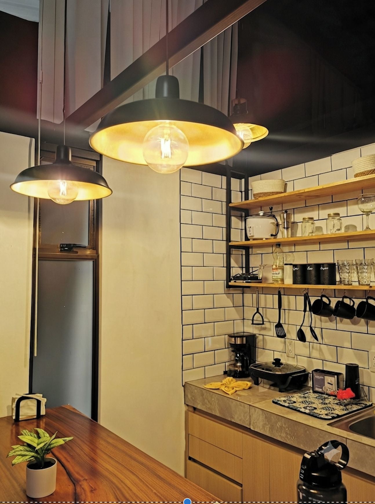 Una cucina moderna con illuminazione a sospensione, un tavolo in legno, una pianta in vaso e uno schienale piastrellato.