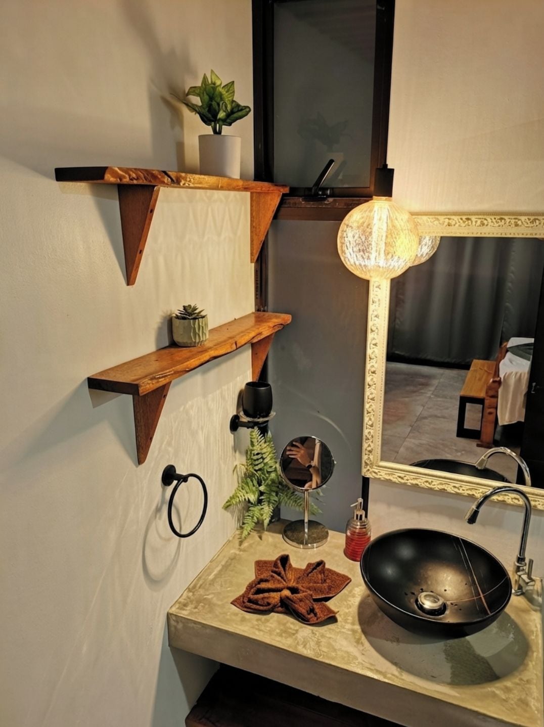 A bathroom with a black sink, a round mirror with a gold frame, and wooden shelves with plants.