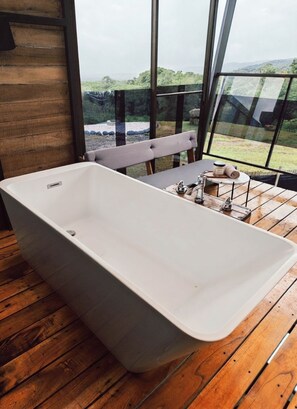 Un baño moderno con una bañera independiente, un banco de madera y vistas a un paisaje natural a través de amplios ventanales.