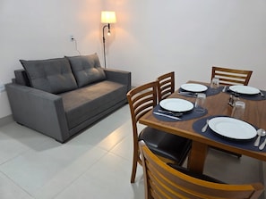 A dining area with a table set for four, a grey sofa, and a wall-mounted lamp. - Studios Flórida (Palmeiras de Goiás)