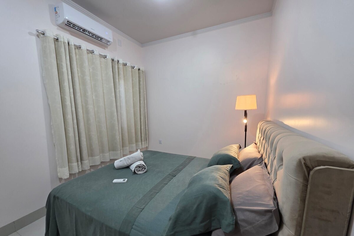 A single bed with a green bedspread, a bedside lamp, and a rolled towel on the bed.