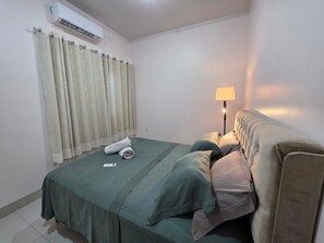 A single bed with a green bedspread, a bedside lamp, and a rolled towel on the bed.