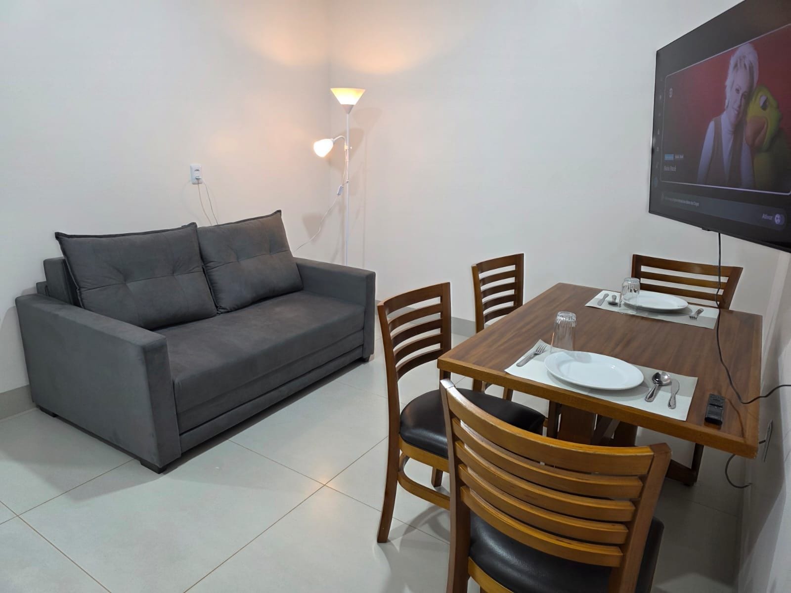 Sala de estar com sofá, mesa de jantar, cadeiras e TV fixada na parede.