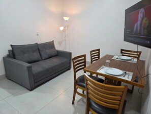 A living area with a sofa, dining table, chairs, and a TV mounted on the wall.
