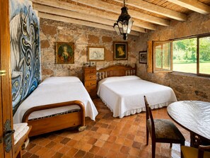 A room with two beds, a wooden dresser, a dining table with a chair, and a large window with a view of greenery.