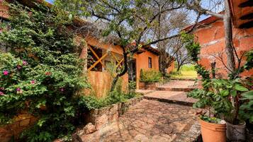 Un patio con senderos de piedra, un pequeño edificio con ventana de cruce y plantas en macetas.