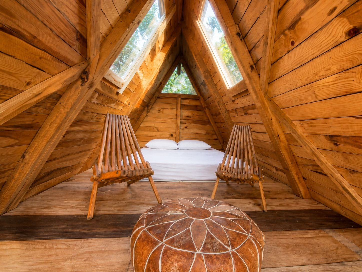 Una habitación de madera con techo inclinado, dos sillas de madera, una cama con ropa blanca y un taburete de madera decorativo.