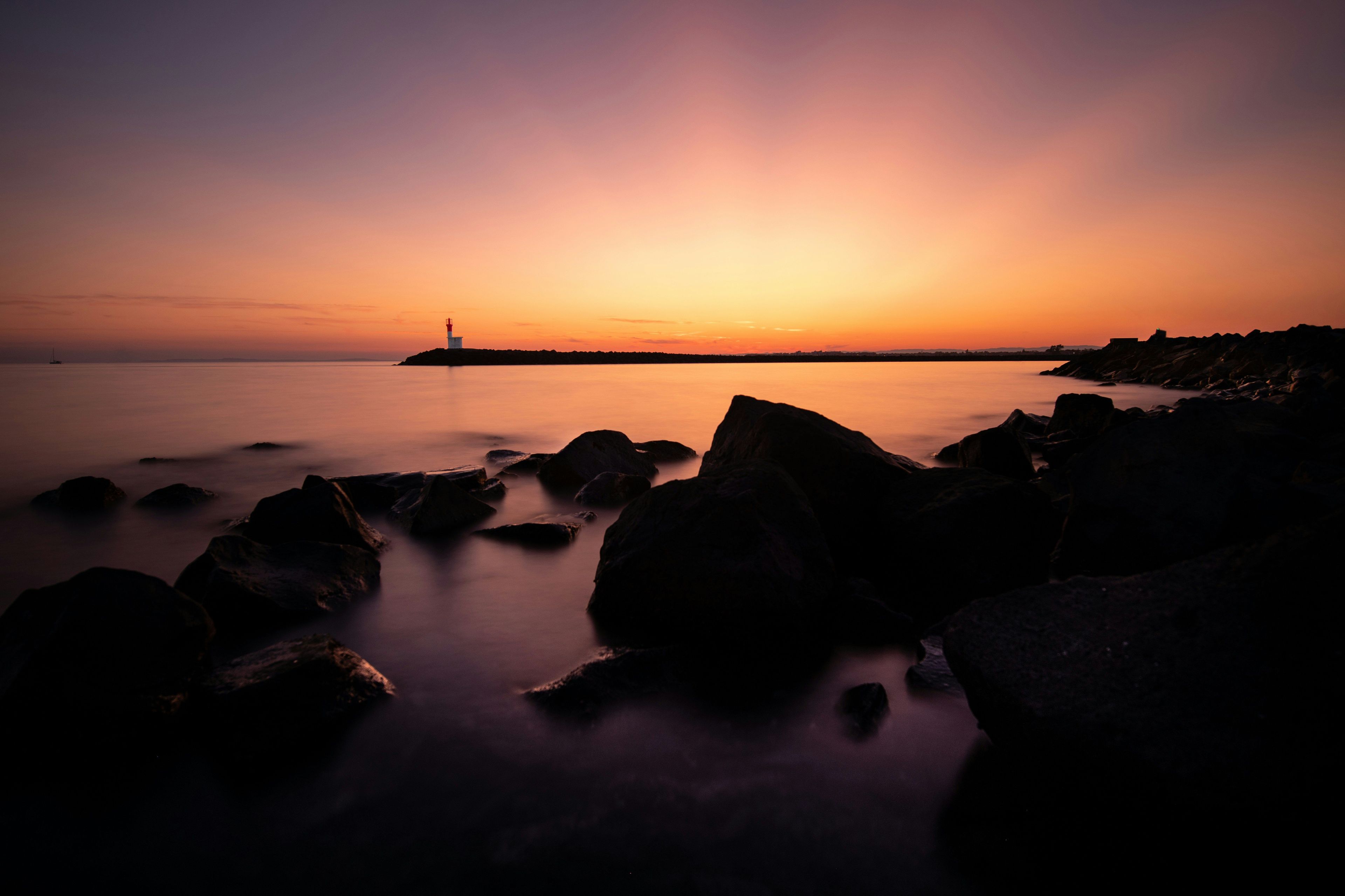 Tramonto sulla costa con la silhouette di un faro, mare calmo e costa rocciosa.